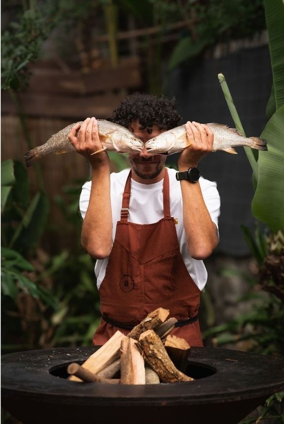 Le chef du Restaurant du Lavoir présentant deux poissons de la pêche locale devant le foyer à bois, au milieu de la végétation.