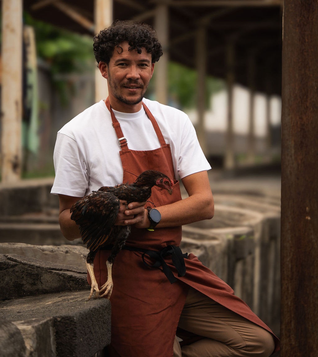 Le chef Matthias Joma, en tablier terracotta, un coq péi dans les bras, dans le quartier de Casabona à Saint-Pierre.