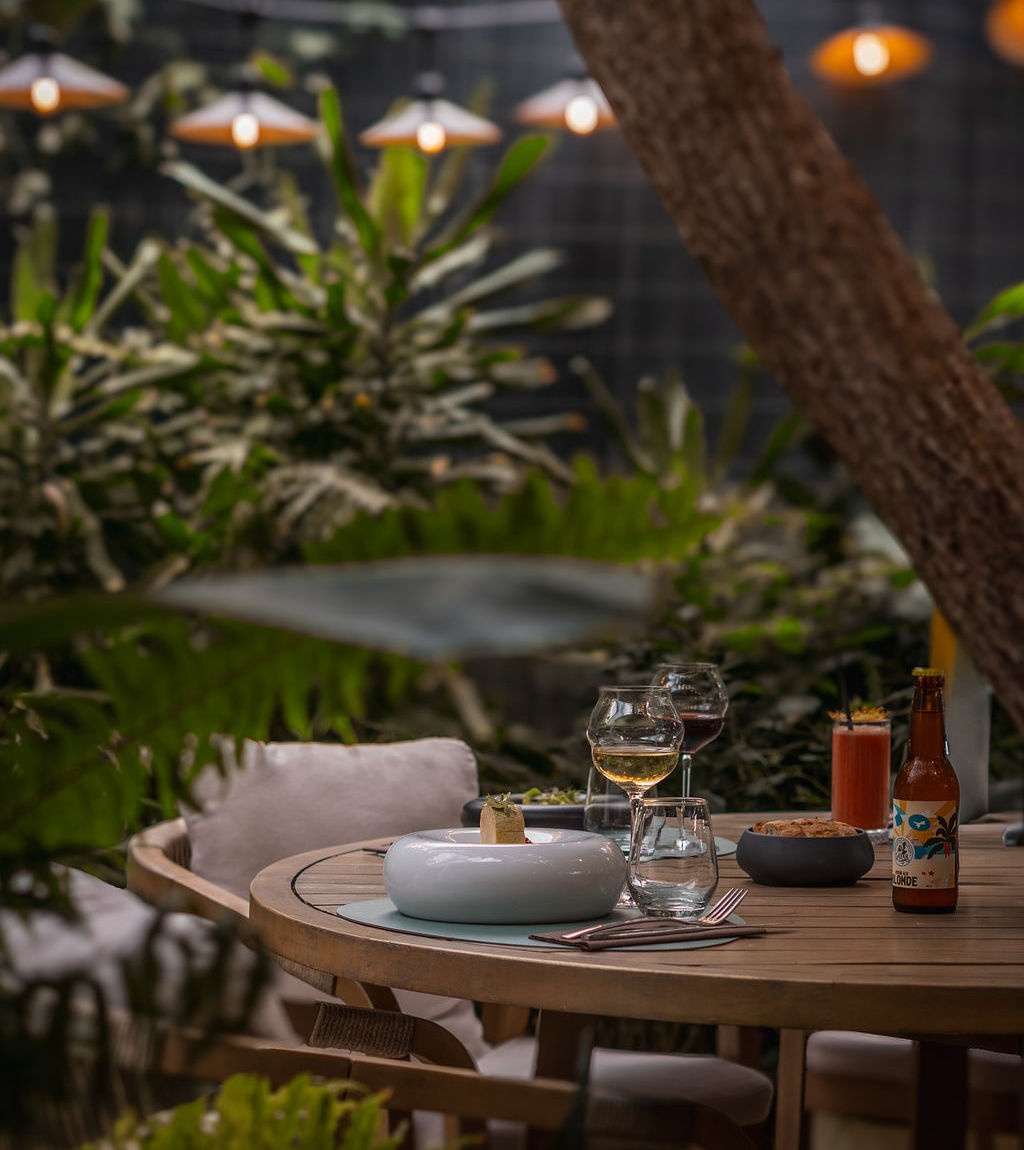 Une table dressée en terrasse au Restaurant du Lavoir, à la tombée du soir, sous les guirlandes lumineuses et la végétation tropicale.